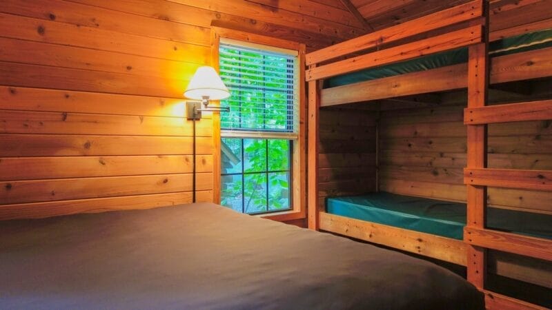 A small wooden cabin bedroom with a single bed, a set of bunk beds, a window with blinds, and a wall-mounted lamp.