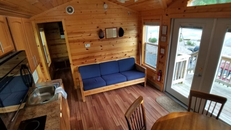 Interior of a small wooden cabin with a blue sofa, kitchen area, round dining table, and glass door leading to an outdoor deck.