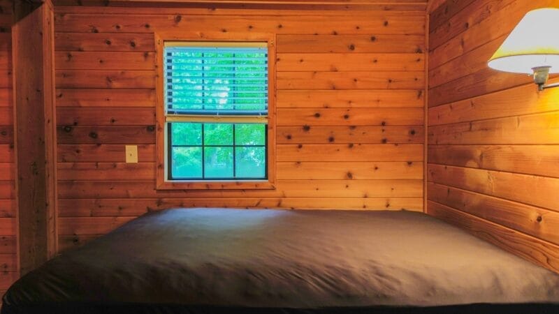 A bed with a dark cover is placed against a wooden wall in a cabin room, with a window and wall-mounted lamp visible.