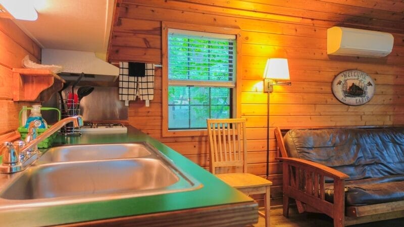 Interior of a wooden cabin with a kitchen sink, counter, stove, window, wooden chair, sofa, wall lamp, and an air conditioner on the wall.