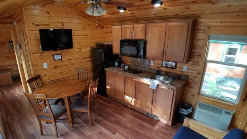 A small wooden cabin interior with a kitchenette, round dining table with four chairs, wall-mounted TV, microwave, fridge, and window with air conditioner visible.