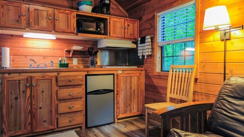 Small wooden kitchen with upper and lower cabinets, a mini fridge, microwave, sink, chair, window with blinds, and a floor lamp, all with wood-paneled walls and flooring.