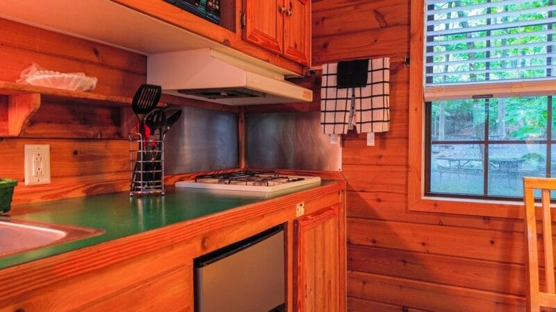 A compact wooden kitchen with a green countertop, a gas stove, utensils in a holder, a checked towel hanging, and a window with blinds showing an outdoor scene.