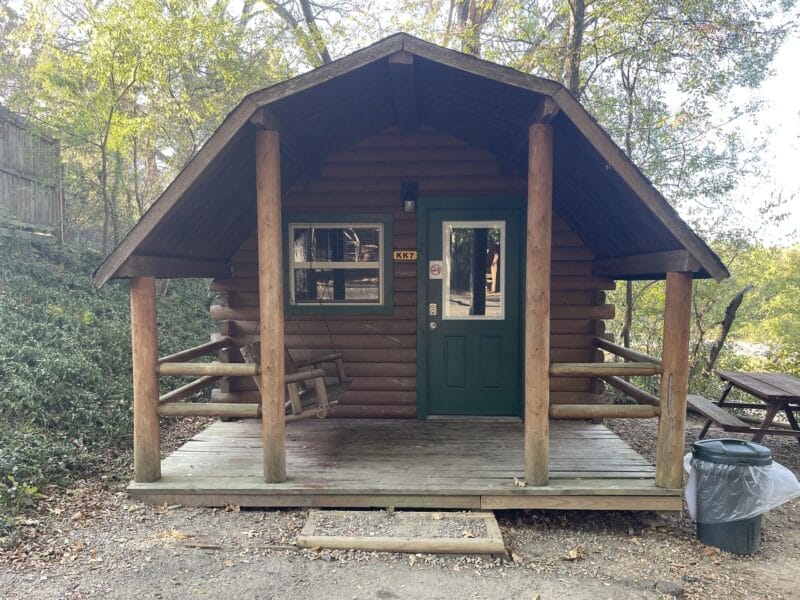 A small log cabin with a green door and porch swing, surrounded by trees. A picnic table and trash can are nearby.