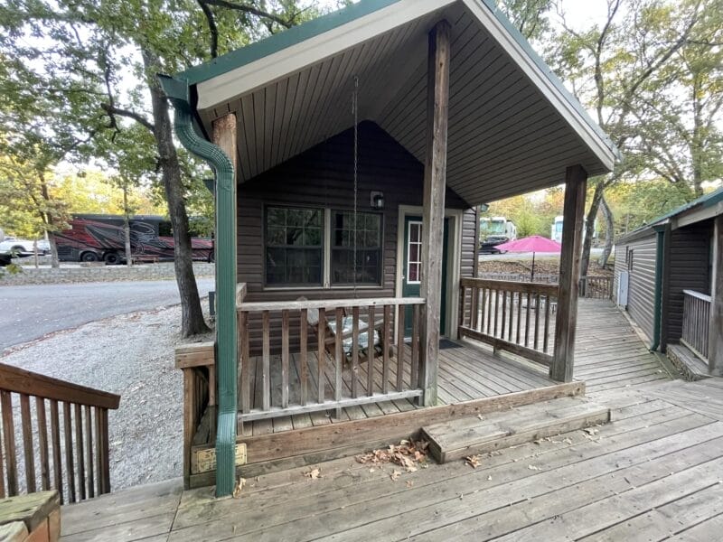 A small wooden cabin with a covered porch and swing, located near a road with parked vehicles and surrounded by trees.