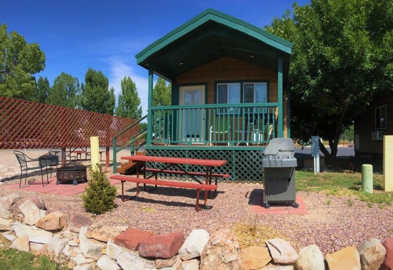 A small cabin with a covered porch, picnic table, grill, fire pit, and outdoor chairs, surrounded by landscaping and trees under a blue sky.