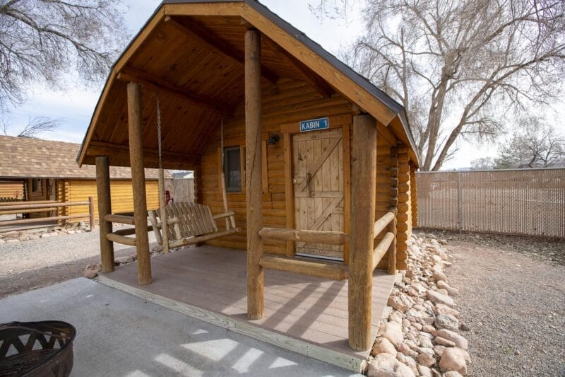 Small wooden cabin with a porch swing, surrounded by rocks, located next to a fence and leafless trees on a clear day.