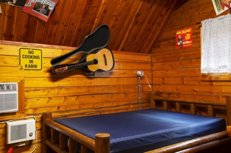 A wooden cabin bedroom with a bare mattress, wall-mounted acoustic guitar in an open case, "No Cooking In Kabin" sign, posters, air conditioner, and a small window with a lace curtain.