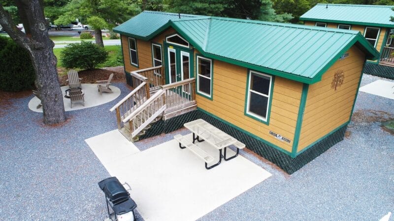 A small cabin with a green metal roof, wooden steps, a picnic table, and a grill sits on a gravel lot surrounded by trees and outdoor seating.