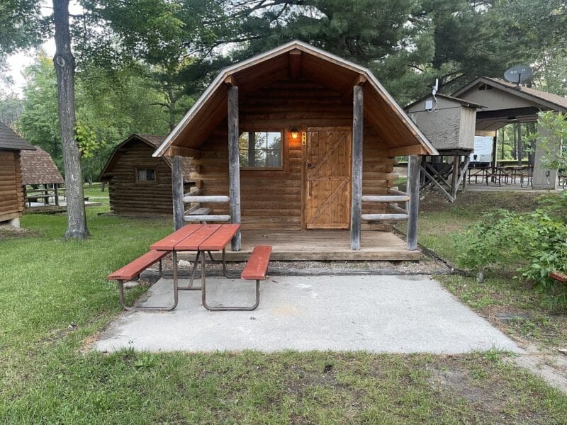 Small log cabin with front porch and lit porch light, set among trees, with a wooden picnic table on a concrete pad in front. Other cabins and greenery are visible in the background.