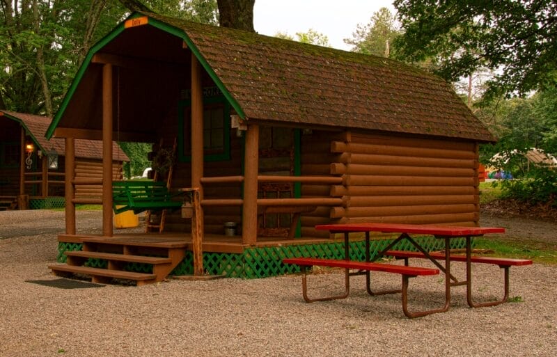 A small log cabin with a covered porch, green swing, and a red picnic table sits on a gravel area surrounded by trees.