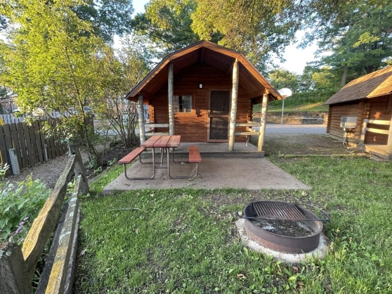 A wooden cabin with a covered porch, picnic table, and fire pit is set on a grassy lot surrounded by trees and a wooden fence.