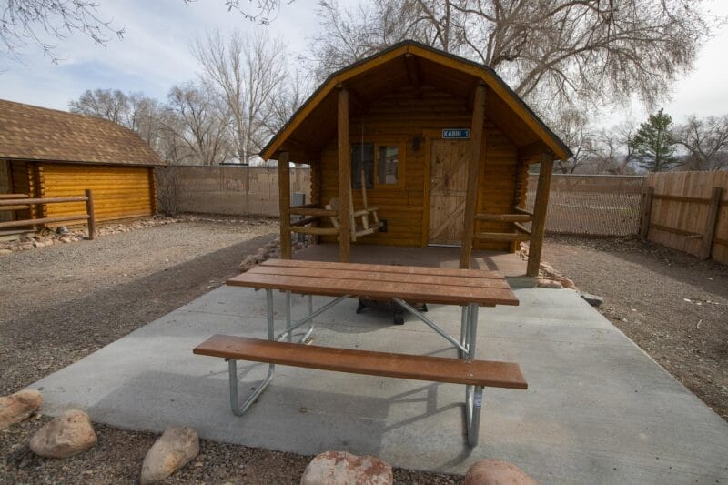A small wooden cabin with a covered porch, flanked by benches, sits behind a picnic table on a concrete pad in a fenced yard with bare trees.