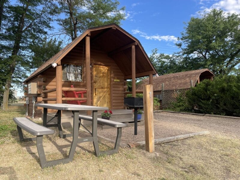 A small wooden cabin with a covered porch, picnic table, and bench sits among trees on a sunny day, with another cabin visible in the background.