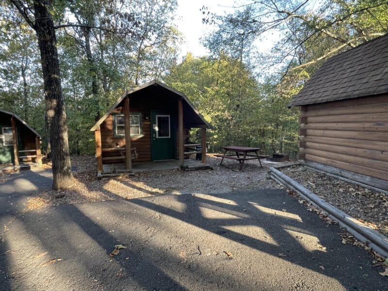 Small wooden cabin with a porch and green door, surrounded by trees. A picnic table and fire ring are nearby. Another similar cabin is partially visible to the right.