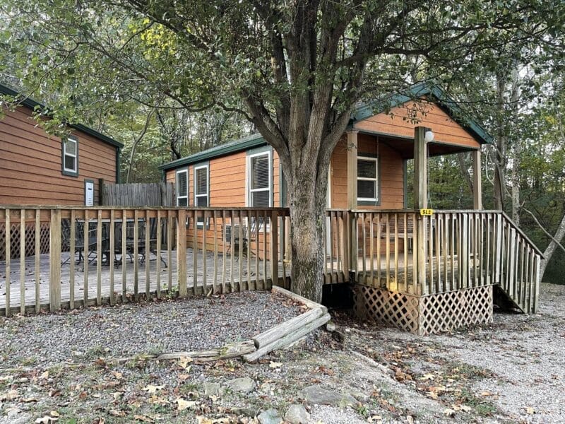 A small wooden cabin with a front porch and ramp, surrounded by trees, with a gravel path and outdoor seating area nearby.