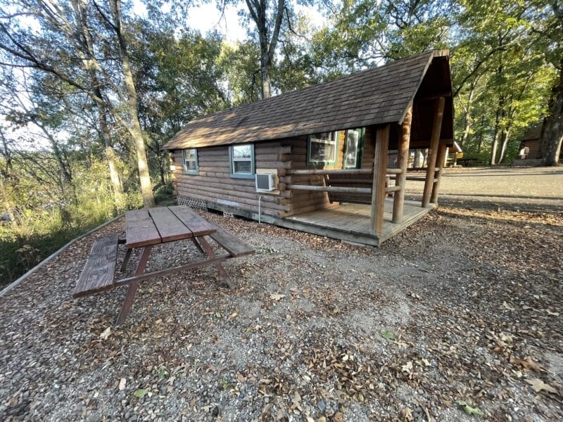 A small wooden cabin with a covered porch sits beside a picnic table on a dirt and gravel area surrounded by trees in daylight.