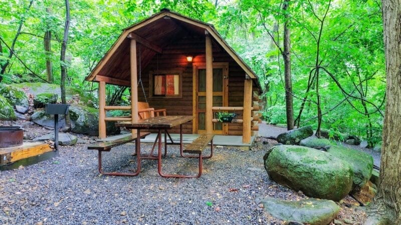 A small wooden cabin with a front porch, picnic table, grill, and campfire ring surrounded by trees and large rocks in a forest setting.