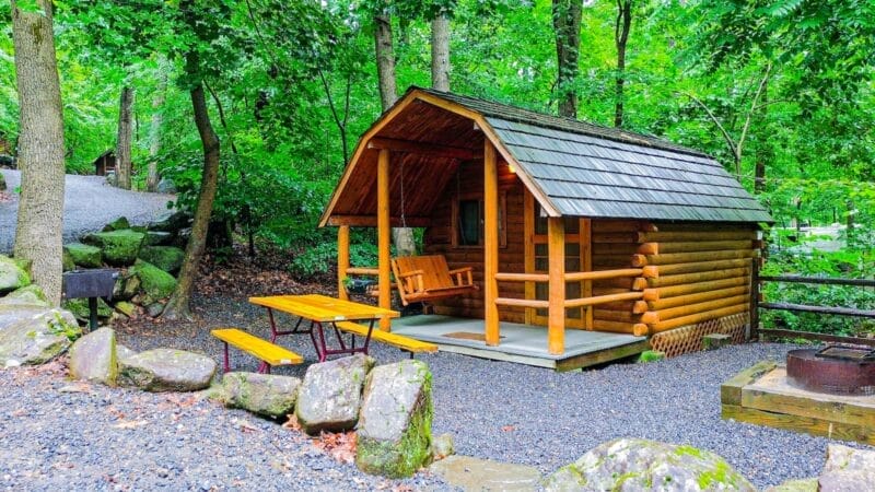 A small wooden log cabin with a front porch, porch swing, picnic table, grill, and fire pit surrounded by trees and rocks in a forested campsite.