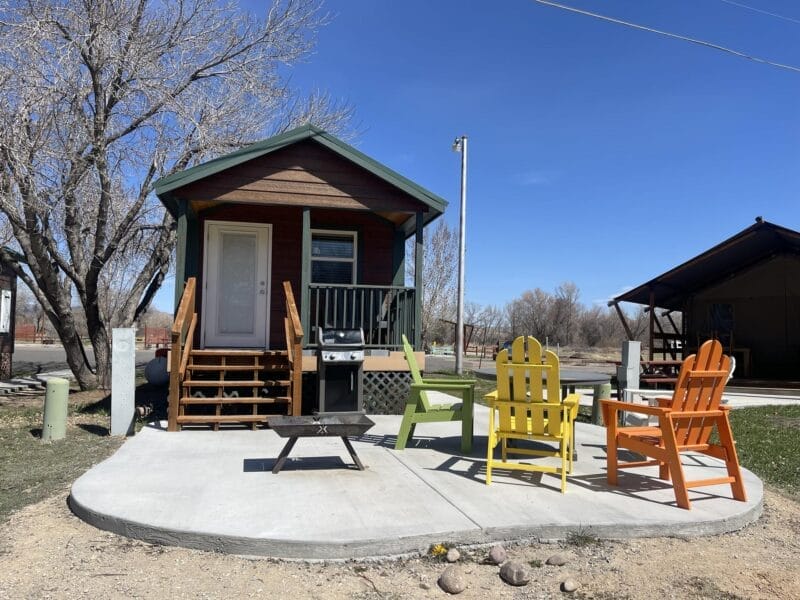 A small wooden cabin with a front porch, barbecue grill, and four colorful Adirondack chairs on a concrete patio, set in a grassy area under a clear blue sky.