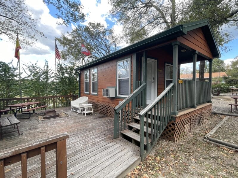 A small wooden cabin with a front porch, outdoor seating, and picnic tables, surrounded by trees and several flags in the background.