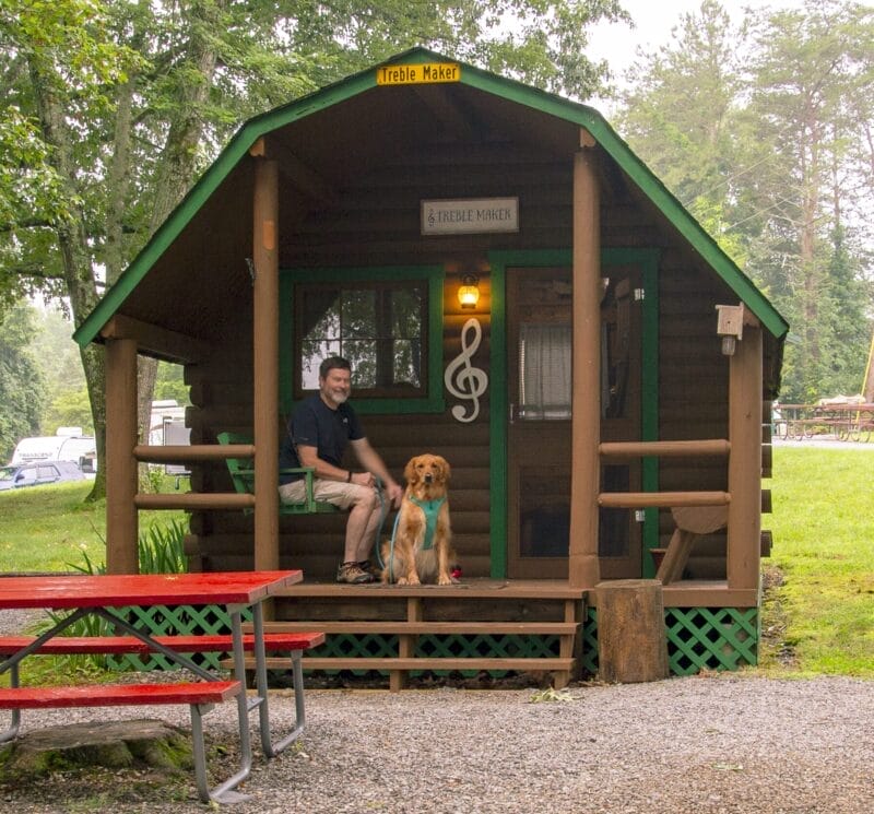A man and a dog sit on the porch of a small wooden cabin labeled "Treble Maker." A red picnic table is in the foreground, and trees are in the background.