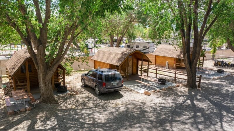 Three wooden cabins with porches are lined up under trees, with a parked SUV and picnic tables nearby. Several RVs are visible in the background behind a fence.