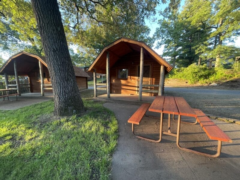 Two small wooden cabins sit near a paved path with a red picnic table under trees on a sunny day.