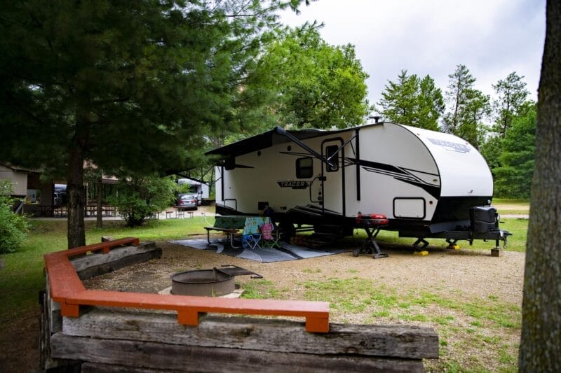 A white travel trailer parked at a campsite with a picnic table, two lawn chairs, a fire pit, and trees in the background.