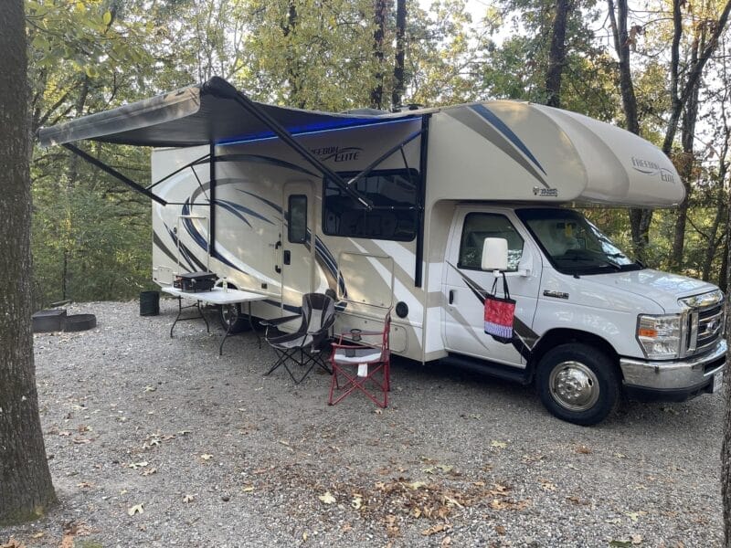 A white motorhome is parked on a gravel campsite with its awning extended, outdoor chairs and table set up, surrounded by trees.