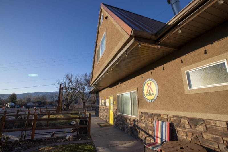 A KOA campground building with brown walls, a sign on the exterior, a chair and table outside, and a clear sky with mountains in the background.