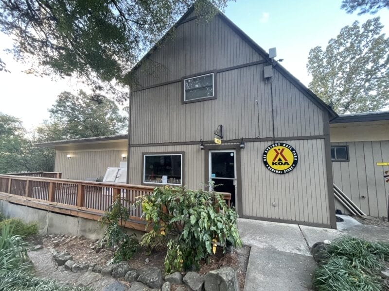 A gray wooden building with a ramp, large windows, and a KOA sign by the entrance, surrounded by greenery and trees.