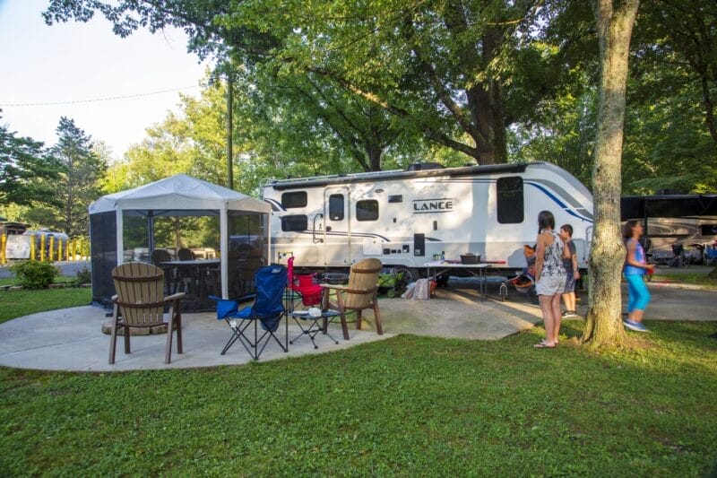 A campsite with a white RV, canopy tent, several chairs, and two people standing nearby under trees on a sunny day.