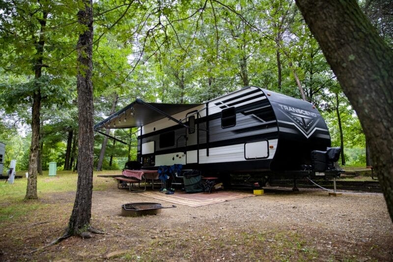 A black and white travel trailer is parked at a wooded campsite with an awning extended, outdoor chairs, and a picnic table nearby.