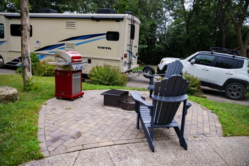 Two Adirondack chairs face a fire pit and red grill on a patio, with an RV and SUV parked nearby in a wooded area.
