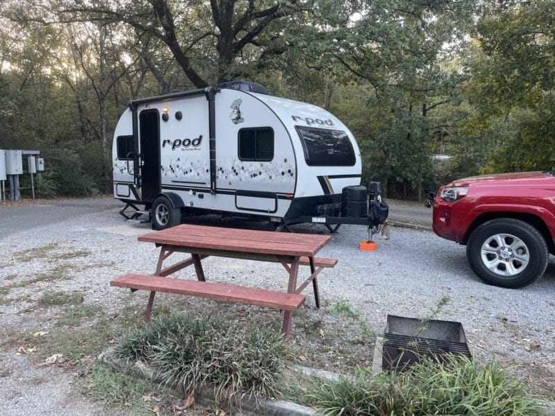 A small travel trailer is parked next to a red vehicle in a gravel campsite with a wooden picnic table and a fire pit, surrounded by trees.