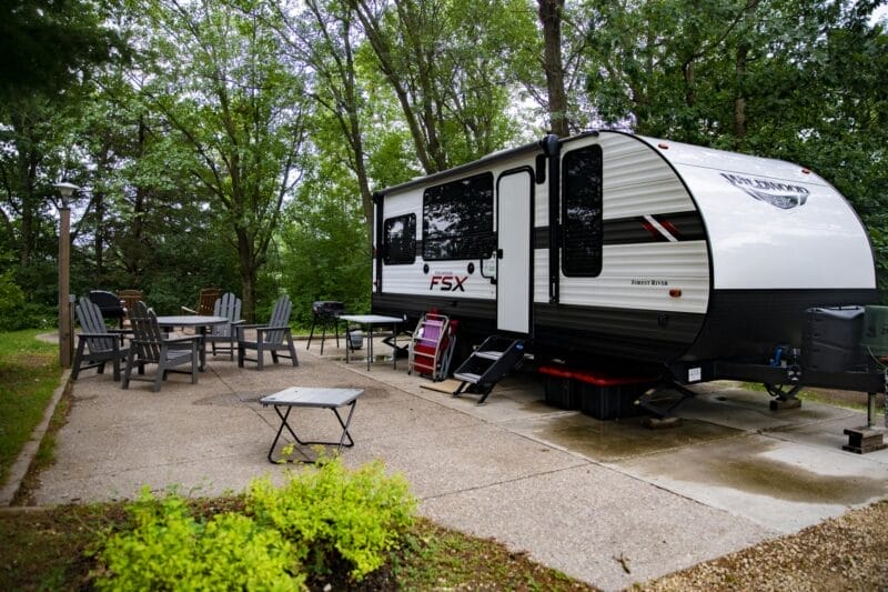 A travel trailer is parked on a concrete pad at a campsite, surrounded by patio chairs, tables, and trees in the background.