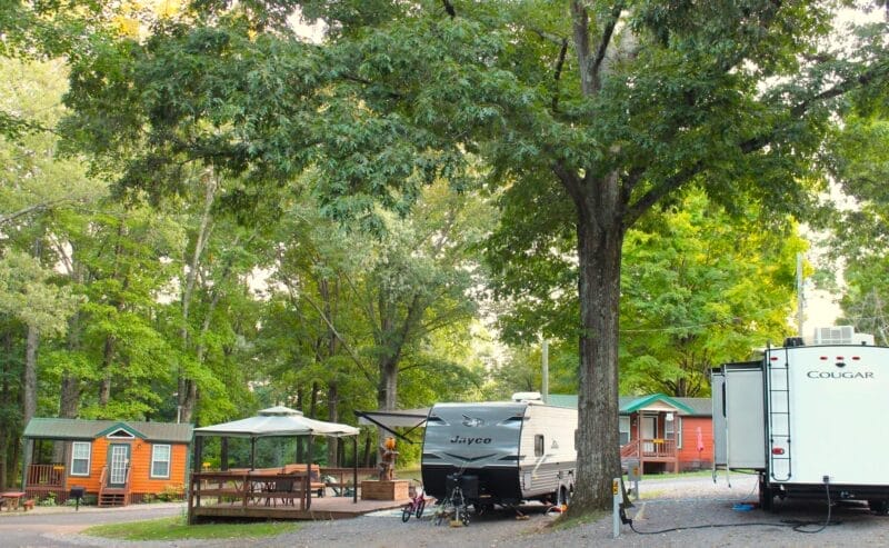 A shaded campground with cabins, picnic tables under a gazebo, and several parked RVs surrounded by tall trees and greenery.