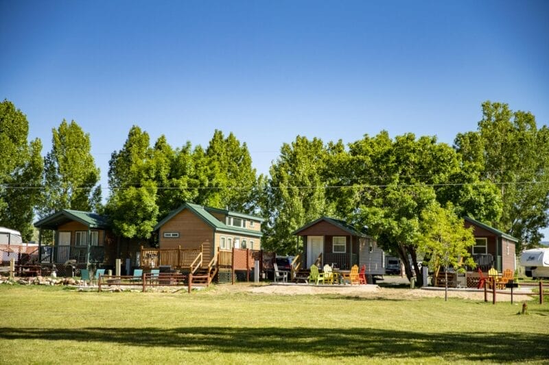 Three small wooden cabins with green roofs sit among trees on a grassy field, with outdoor chairs and a clear blue sky in the background.