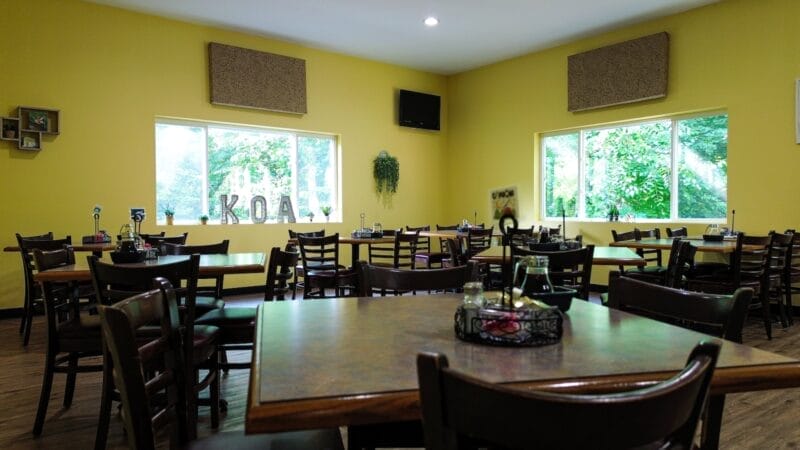 A dining area with several empty wooden tables and chairs, yellow walls, large windows letting in natural light, and a small wall-mounted TV.
