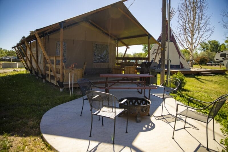 A glamping tent with a shaded porch, outdoor seating area with metal chairs and a fire pit, set on a concrete pad in a grassy campground.
