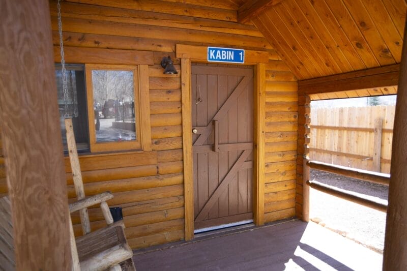 Wooden porch with a rustic bench, window, and a brown door labeled "KABIN 1" on a blue sign; wooden fence and trees visible in the background.