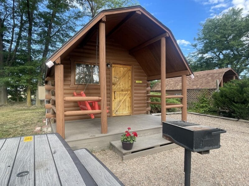 A small log cabin with a porch and red chair sits beside a picnic table and grill, surrounded by trees and a gravel yard under a partly cloudy sky.
