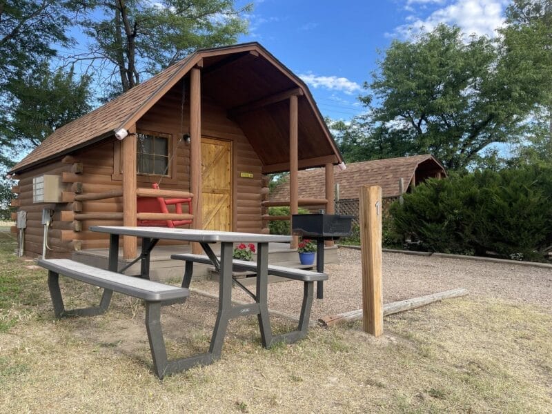 A small log cabin with a porch, picnic table, and grill sits on a gravel lot surrounded by trees and shrubbery under a partly cloudy sky.
