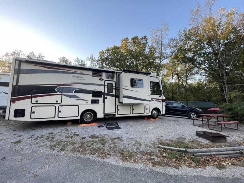 A large RV and a black car are parked on a gravel lot next to a picnic table, grill, and some trees at a campsite.