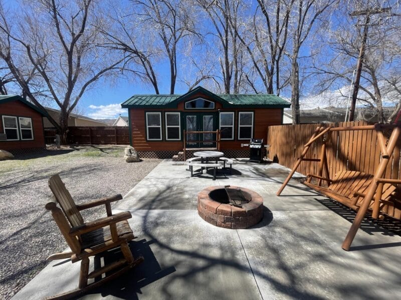 A cabin with green roof and multiple windows sits behind a patio with a fire pit, wooden chairs, a swing, and a grill, surrounded by leafless trees.