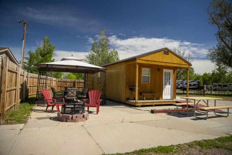 A small yellow cabin with a white door stands on a concrete lot, next to a gazebo with red chairs, a fire pit, and a picnic table. RVs are visible in the background.