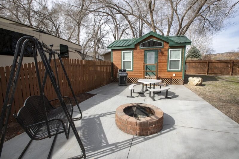 A small wooden cabin with a green metal roof sits behind a fenced patio with a metal swing, a round picnic table, and a brick fire pit. Leafless trees are in the background.