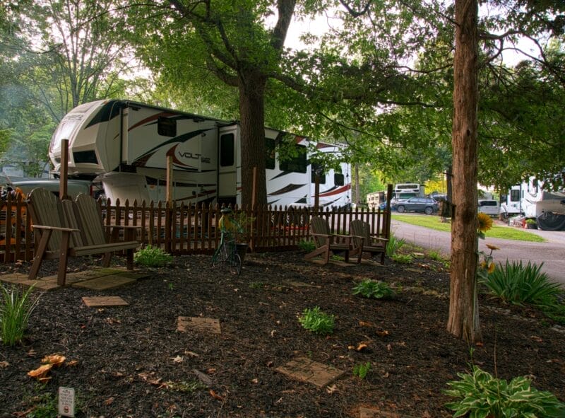 A fenced garden area with wooden chairs and plants sits in front of several large RVs parked under tall trees at a campground.