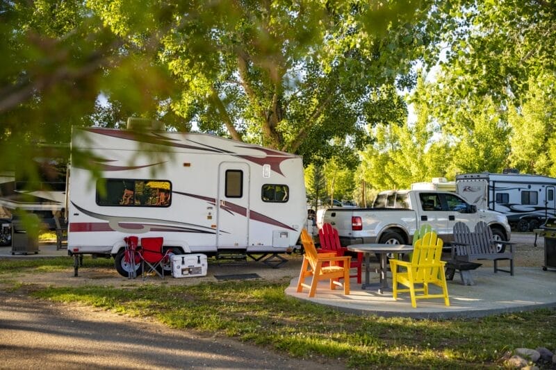 A white camper trailer parked at a campsite with colorful Adirondack chairs and a table nearby, surrounded by trees and other RVs.